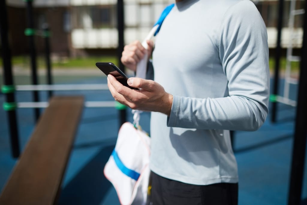 Urheilulliseen pitkähihaiseen paitaan pukeutunut mies katsoo mustaa älypuhelinta ulkona.