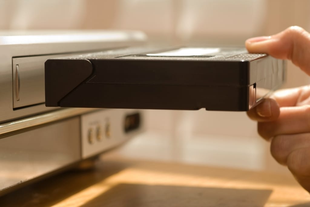 Käsi työntää VHS-videokasettia vanhaan videonauhuriin.