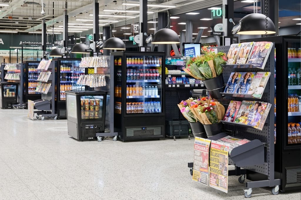 Ruokakaupan kassa-alue, jonka hyllyillä on virvoitusjuomia, lehtiä ja kukkakimppuja.