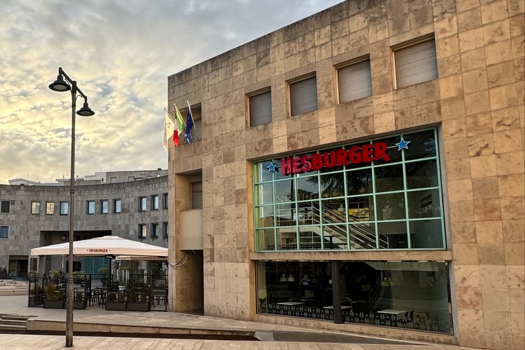 Hesburgerin ravintolan kivijulkisivu, suuri lasiseinä ja ulkoterassi Italian Materassa päivänvalossa.
